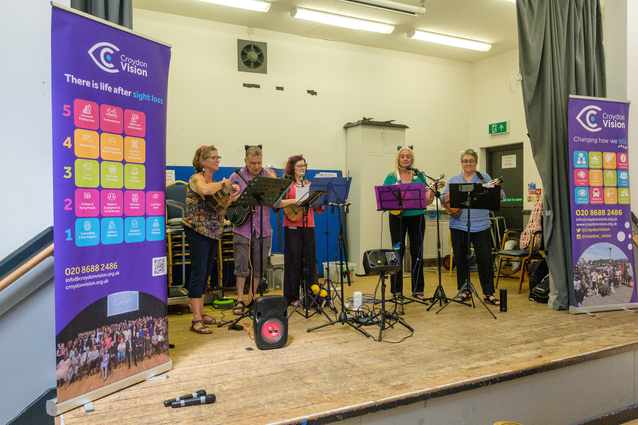The Wednesday Warblers performing with ukuleles and microphones between two Croydon Vision banners.