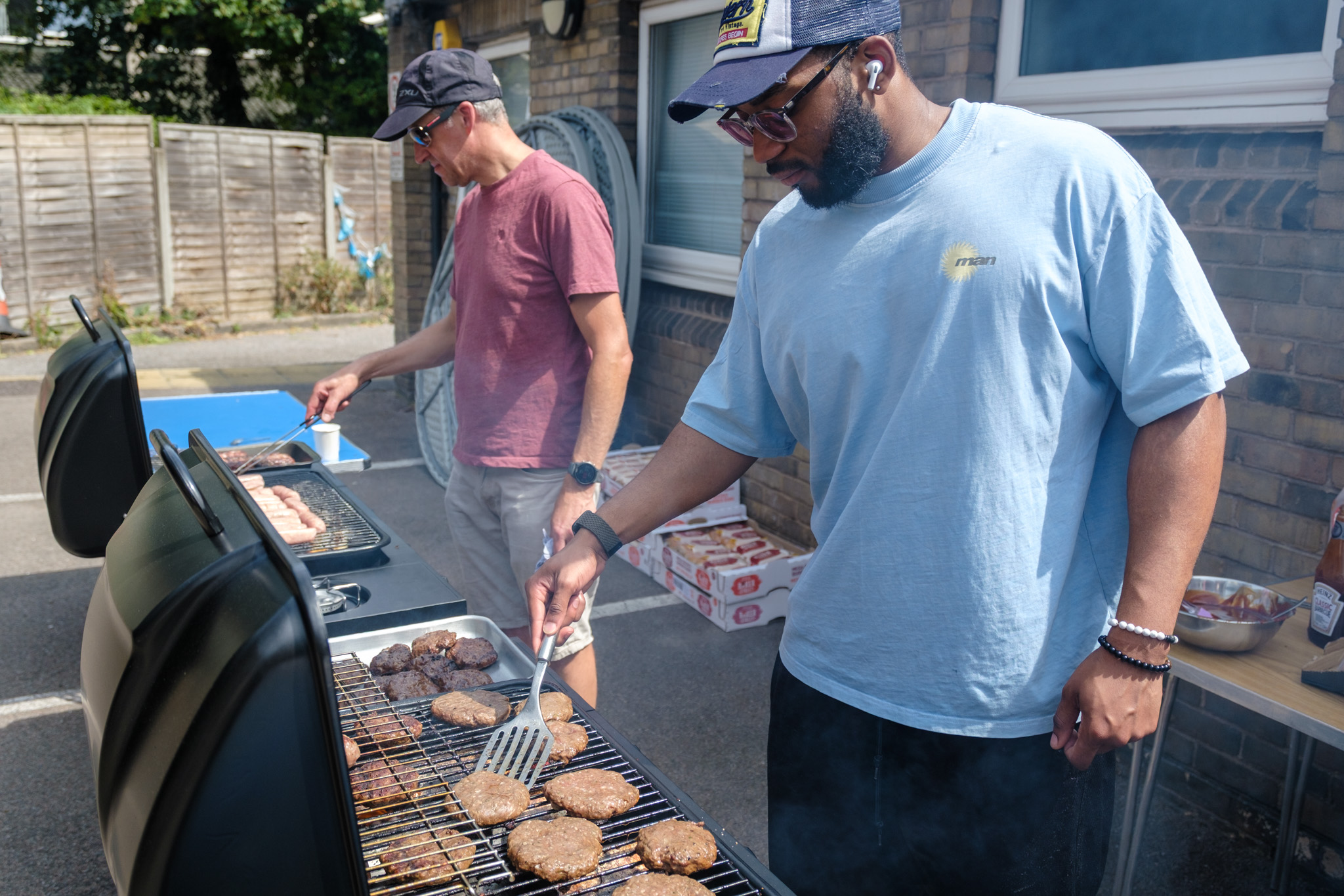 Two people cooking burgers and sausages on outdoor barbecues.